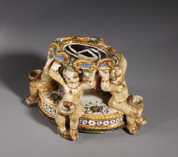 BOTTEGA DEI PATANAZZI, URBINO, FINE DEL XVI SECOLO : Saliera in maiolica policroma poggiante su base ovale su cui si trovano quattro putti seduti che sorreggono l'invaso superiore, decorato nell'incavo da una figura allegorica in grisaille  - Asta Asta 387 | ARTE ANTICA E DEL XIX SECOLO - DAL RINASCIMENTO AL SIMBOLISMO: OPERE DA COLLEZIONI ITALIANE Tradizionale - Associazione Nazionale - Case d'Asta italiane