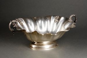 ARGENTIERE ITALIANO DEL XX SECOLO - Alzata centrotavola in argento con bordo mistilineo e decorazioni vegetali a rilievo, manici in forma di delfini, poggiante su base ovale