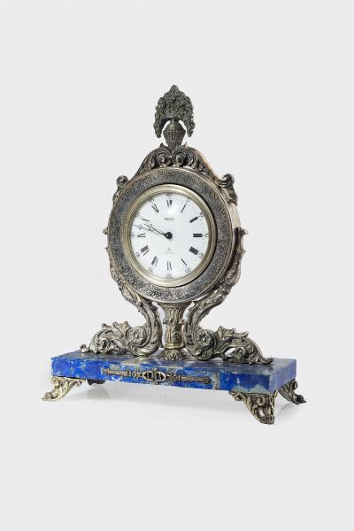 <BR>23x17 cm<BR>Cassa in argento decorata con racemi vegetali e vaso con fiori. Quadrante in smalto con numeri romani serigrafati e minuteria periferica a binario.<BR>Poggiante su base in lapislazzuli e quattro piedini sbalzati.  - Asta Orologi  - Associazione Nazionale - Case d'Asta italiane