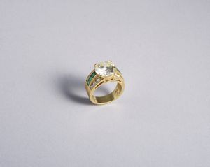. - Anello in oro giallo 18k con diamante centrale taglio a brillante di 6,32 carati (vedi certificato IGI ) con 12 diamanti bianchi ovali di 1,20 ct. totali e 6 smeraldi taglio carr.