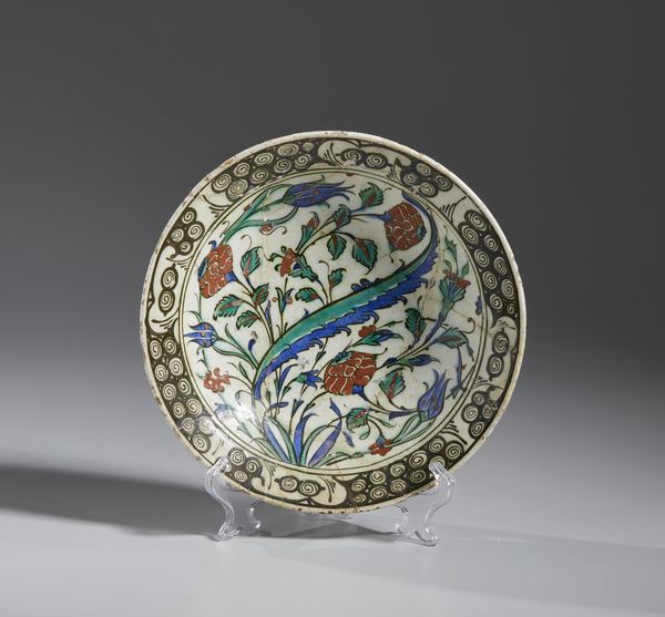 MANIFATTURA DI IZNIK, XVII SECOLO : Piatto in ceramica invetriata policroma con cavetto centrato da una foglia lanceolata attorniata da garofani rossi, tulipani blu e tralci fogliati, tesa decorata con motivo 'ad onde e scogli'  - Asta Asta 432 | ARTE ANTICA E DEL XIX SECOLO Tradizionale - Associazione Nazionale - Case d'Asta italiane