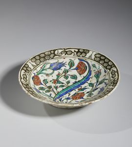 MANIFATTURA DI IZNIK, XVII SECOLO : Piatto in ceramica invetriata policroma con cavetto centrato da una foglia lanceolata attorniata da garofani rossi, tulipani blu e tralci fogliati, tesa decorata con motivo 'ad onde e scogli'  - Asta Asta 432 | ARTE ANTICA E DEL XIX SECOLO Tradizionale - Associazione Nazionale - Case d'Asta italiane